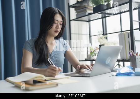 Young Asian Woman Using Laptop for Work at Home on Modern Desk Setup in Cozy blue House Stock Photo