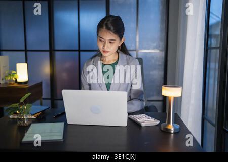 Alone Working Late at Night. Asian business woman at workplace in office late night work at a desk in a dark office working on a laptop late in the ev Stock Photo