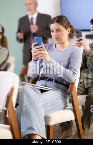 Young female student scans QR code or takes photo, teacher in background Stock Photo