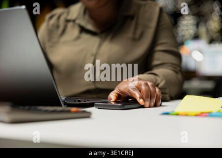 African american worker checking her mobile phone agenda before scheduling appointment, picking up phone to read her notifications. Woman getting distracted at work after hours. Stock Photo