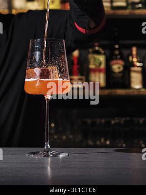 Mixologist expertly pours a vibrant orange cocktail into a glass against a backdrop of shelves stocked with various bottles Stock Photo
