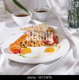 Full english breakfast with fried eggs, sausages, beans, and toasts is served on white tablecloth Stock Photo