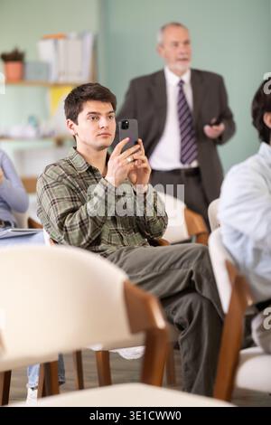 Young male student scans QR code or takes photo, teacher in background Stock Photo