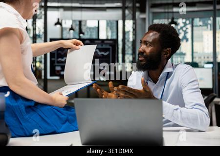 Black man and female coworker reviewing analytics, paperwork and reports at modern office desk. Multiracial business partners working on project planning, financial tasks and investment strategies. Stock Photo