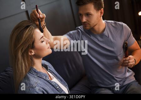 Young bearded man sitting on sofa is applying makeup to a beautiful young woman in a dimly lit room Stock Photo