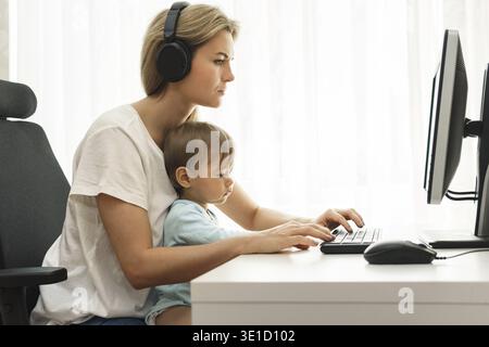 Young freelancer mother is working on computer at her home office workplace with her little son sitting on her lap Stock Photo