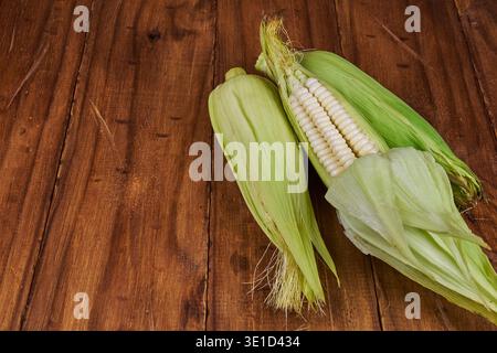 White corn cobs with husks providing fresh, organic farm produce on a natural wood surface Stock Photo
