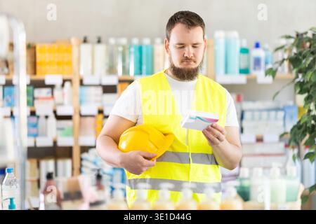 Male builder in yellow vest with helmet in his hands looks for medicine in pharmacy Stock Photo
