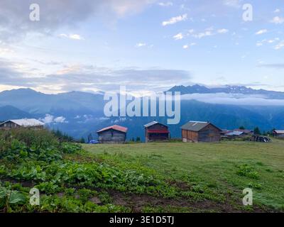Traditional wooden farm buildings sit on a green hillside meadow overlooking a spectacular mountain valley. Dramatic clouds drift through the alpine p Stock Photo