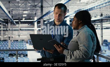 Team of engineers monitor server hub activity using laptop and tablet. Teamworking data center colleagues review rigs tests results on devices to ensure systems are working correctly, camera B Stock Photo