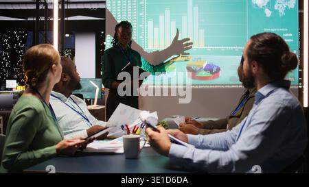 Black team leader from a corporation discussing financial details and insights in a new project presentation during late night business meeting. Shareholders share development ideas. Camera A. Stock Photo