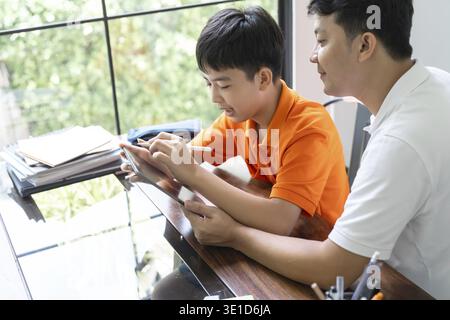 Asian father and son using tablet teaching son to draw playing with digital tablet at desk fatherhood concept Stock Photo