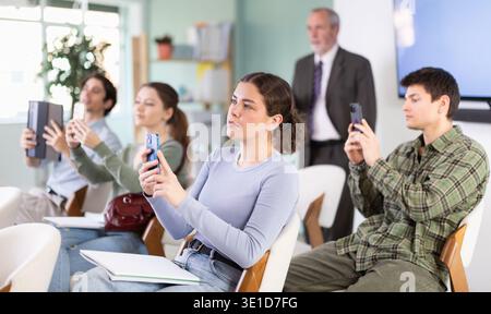 Young female student scans QR code or takes photo, teacher in background Stock Photo
