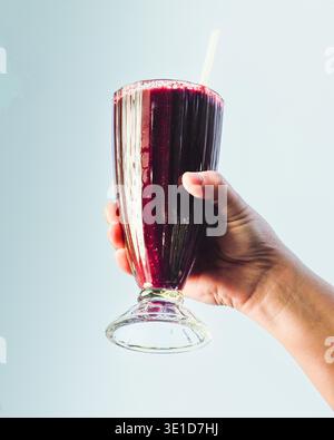 Hand holds tall glass filled with deep purple smoothie. Glass features a white straw and clear stem base. Bright light highlights the vibrant, rich co Stock Photo