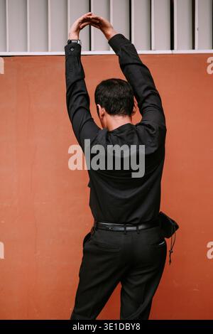 A person in dark attire is captured from behind, stretching their arms overhead against a warm orange wall. The simple background highlights the actio Stock Photo