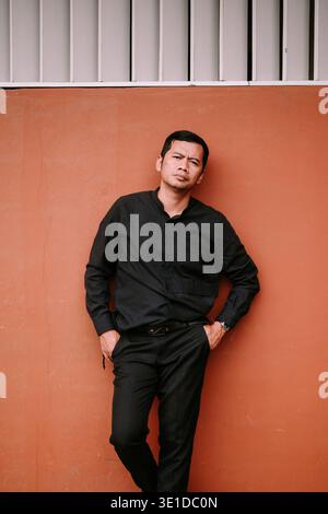 A stylish man in a black collared shirt and pants stands confidently with hands in pockets, leaning against a textured orange wall outdoors. Stock Photo
