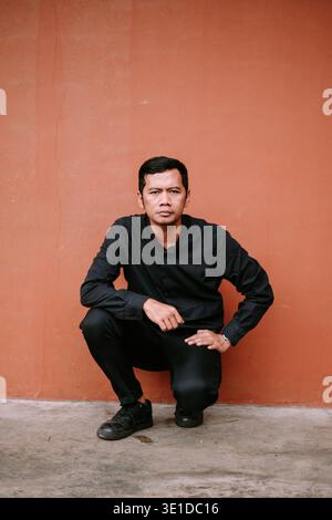 A focused young man dressed in all black squats confidently in front of a warm orange wall. His intense gaze conveys determination and strength, suita Stock Photo