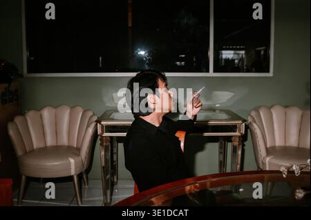 A contemplative young man sits indoors, smoking a cigarette with visible smoke swirling. He is focused, perhaps taking a moment to reflect in the dim Stock Photo