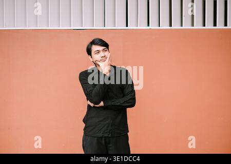 A thoughtful young Asian man stands against a peach-colored wall, hand on chin, looking upward in contemplation. He appears to be deeply engrossed in Stock Photo