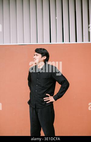 A thoughtful young man in a black shirt stands confidently against a terracotta-colored wall, looking sideways. His casual attire and relaxed pose cre Stock Photo