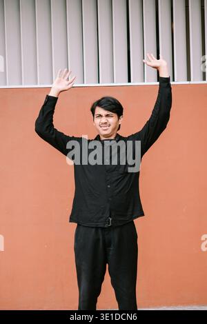 A young Asian man stands against an orange wall, raising both hands in a gesture of frustration and helplessness. His facial expression conveys annoya Stock Photo