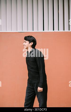 A happy young Asian man in a black shirt stands confidently in profile, smiling against a vibrant orange and white architectural background. Stock Photo