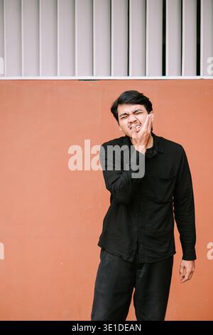 A young man is grimacing and holding his cheek due to intense toothache or jaw pain, standing against an orange wall. He appears to be suffering from Stock Photo