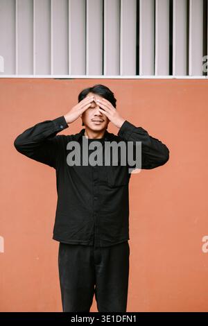 A young man covers his eyes with his hands, displaying feelings of stress, fatigue, or discomfort. He stands against a vibrant orange wall. Stock Photo