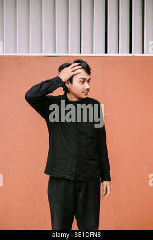 A thoughtful young man in a black shirt poses with his hand on his head against an orange-brown wall, looking sideways. He appears to be contemplating Stock Photo