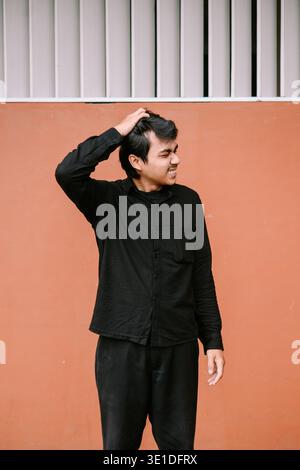 A young man in a black shirt scratches his head with an intense, frustrated expression against a simple orange wall. He conveys feelings of stress, co Stock Photo