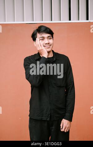 A young Asian man in a black long-sleeved shirt poses against an orange wall, looking thoughtfully to the side with a gentle smile. Stock Photo