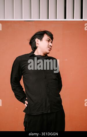 A young adult man dressed in a casual black long-sleeved shirt and dark pants stands with hands on his lower back against a warm orange-brown wall. He Stock Photo