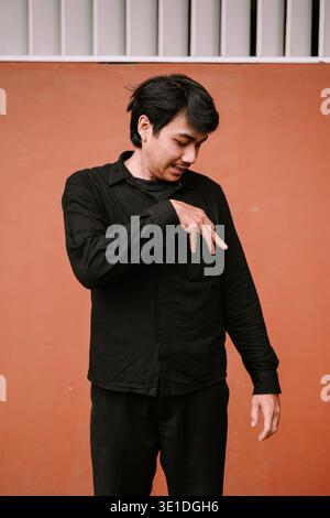 A young Asian man wearing a black shirt stands thoughtfully against a vibrant orange wall, making an expressive hand gesture. He appears pensive and r Stock Photo