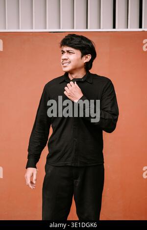 A young Asian man in a black shirt stands against an orange wall, looking perplexed and adjusting his collar. His furrowed brows and slight grimace co Stock Photo