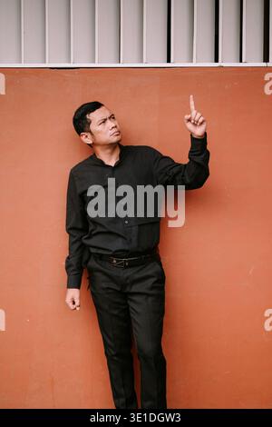 An adult man in a black shirt and pants points his index finger upwards against a vibrant orange-red wall. His focused expression suggests attention, Stock Photo