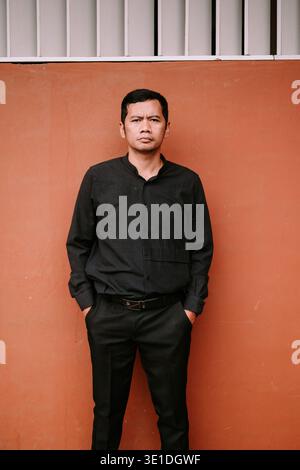A man in a stylish black shirt and pants stands confidently against a terracotta wall, hands in pockets. His serious expression conveys contemplation Stock Photo