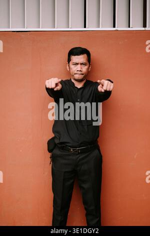 A serious man wearing a black shirt and pants stands against an orange wall, pointing both hands directly towards the viewer with a determined express Stock Photo