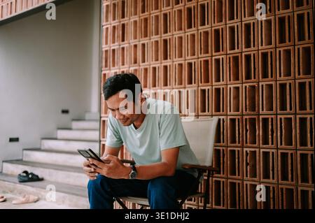 A casual young man smiles while engrossed in his smartphone, sitting on a modern patio. The stylish background with a terracotta brick wall and stairc Stock Photo