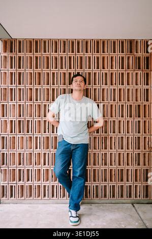 A man in a light t-shirt and jeans stands confidently against a modern textured terracotta brick wall, posing with a neutral expression for a relaxed Stock Photo