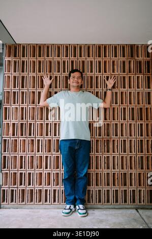 A casual man in a light green t-shirt and blue jeans smiles while posing against a modern terracotta brick wall. His hands are raised against the text Stock Photo