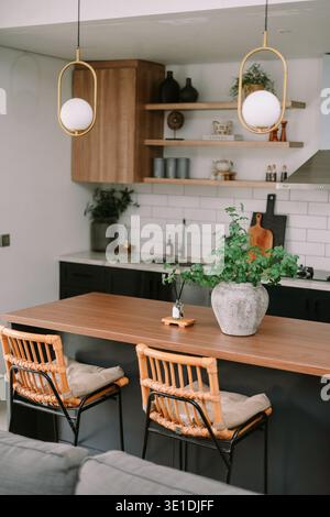 A stylish contemporary kitchen interior featuring a wooden island, rattan stools, and modern pendant lights. Green plants add a fresh touch to this in Stock Photo
