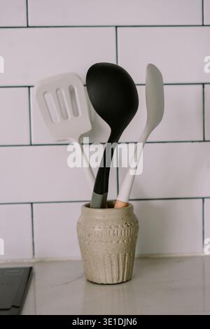 Modern kitchen utensils, including a spatula and ladle, are neatly organized in a stylish textured ceramic holder on a clean counter. White subway til Stock Photo