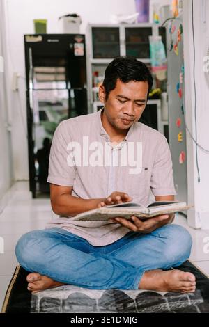 An adult man sits cross-legged on a prayer mat, intently reading from a religious book. He is focused on his scripture in a quiet indoor setting, demo Stock Photo