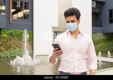 Portrait of young Indian businessman with mask for protection from corona virus outbreak outside modern building in the city Stock Photo
