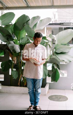 A man stands outdoors, focused on reading a book. He is dressed casually, surrounded by large green plants on a patio or balcony. Stock Photo