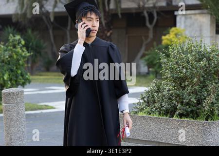 Asian man wearing gown and cap, holding diploma with red ribbon, using smartphone on campus walkway Stock Photo
