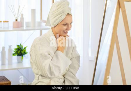 Happy beautiful woman enjoys appearance applying cosmetcs, cream product to face looking in mirror Stock Photo