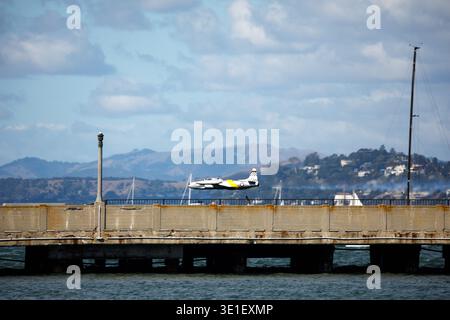 A model airplane flies over a pier with sailboats and hills in the background. Stock Photo