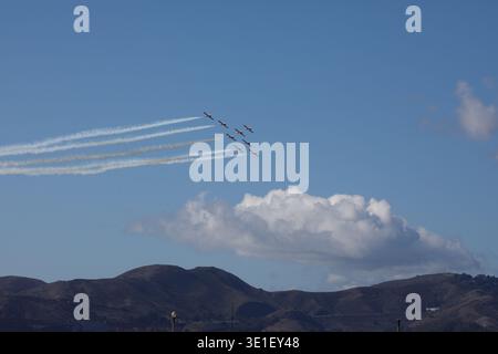 A formation of seven red and white planes flies in formation, leaving white contrails in the blue sky above rolling hills. Stock Photo