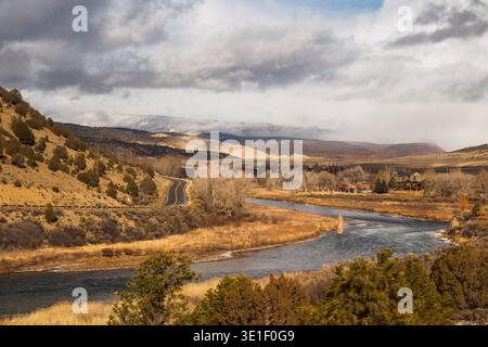 A winding road leads through a valley with a river and snow-capped mountains in the background under a cloudy sky. Stock Photo
